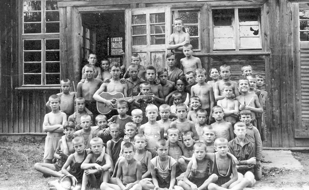zdjęcie czarno-białe przedstawiające grupę dzieci stojących przed drewnianym budynkiem