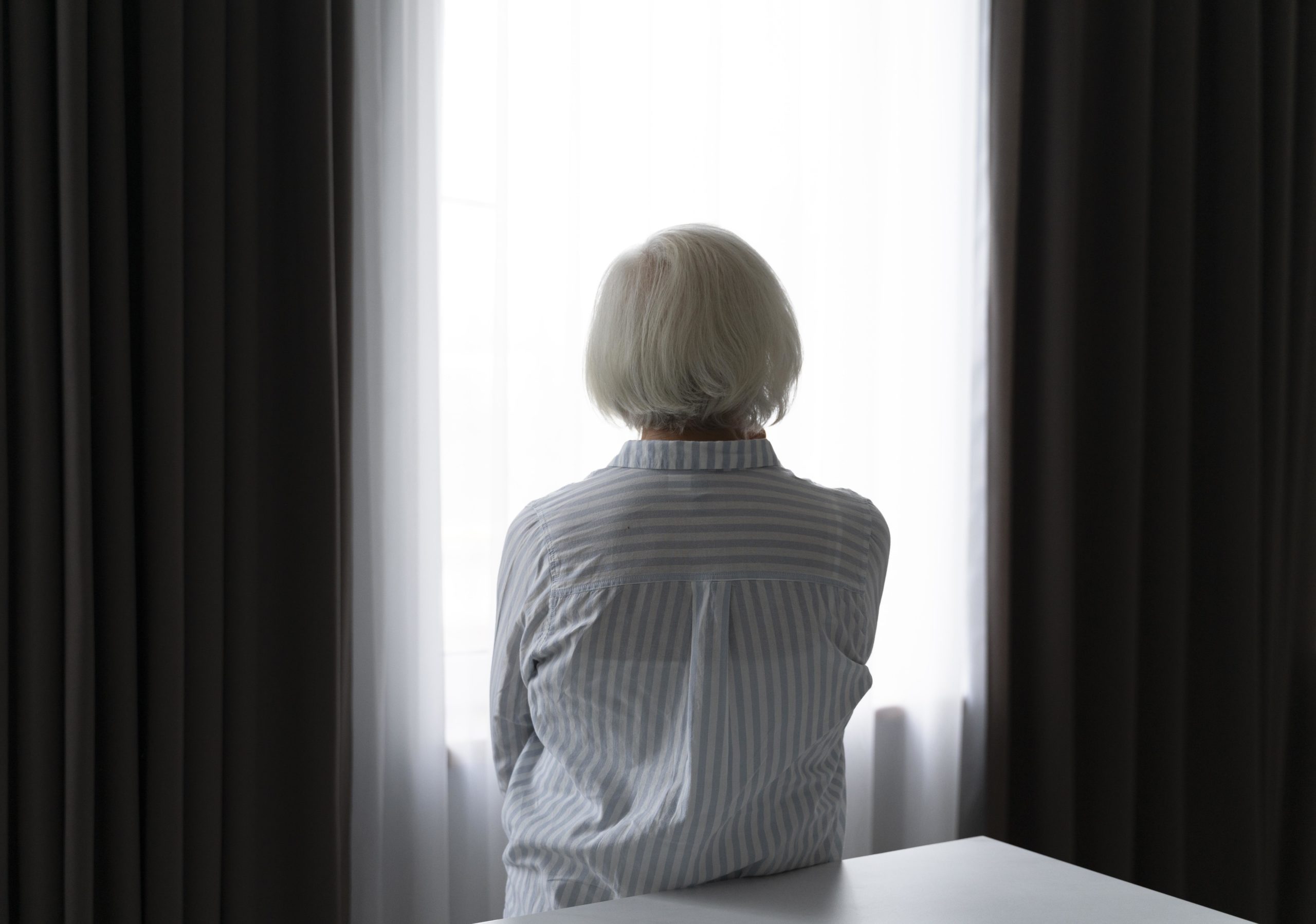 starsza kobieta, stojąca tyłem, patrząca w okno