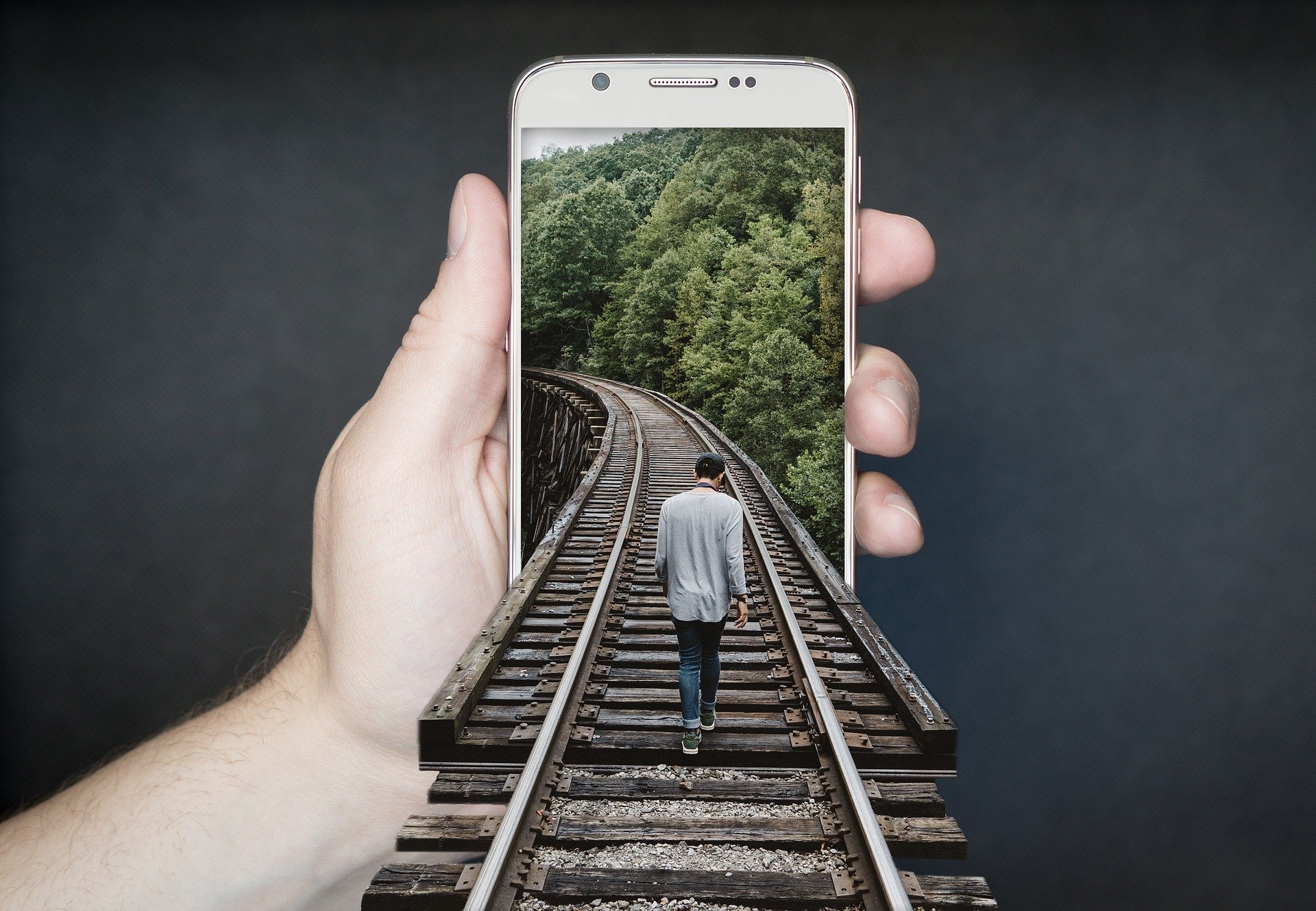 dłoń trzymająca smatfona, na ekranie smartfona mężczyzna idący po torach kolejowych