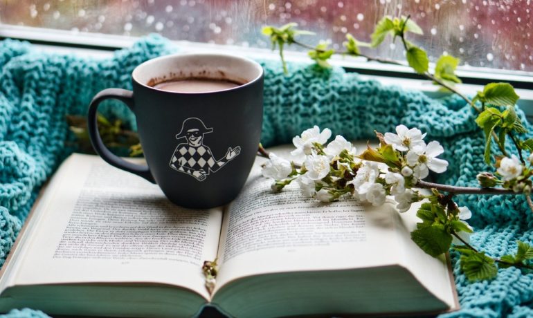 kubek z kawą i kwiaty na książce i wełnianym kocu przy oknie z mokrą szybą