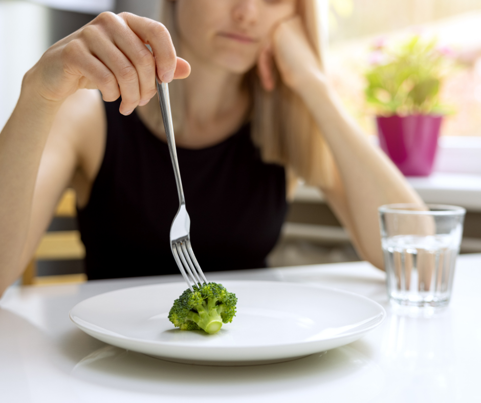 Zdjęcie. Kobieta siedząca za stołem z widelcem w ręku. Na talerzu przed nią leży brokuł.